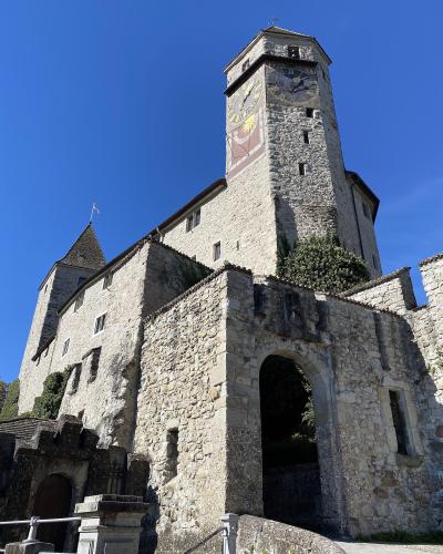 A low-angle view of a medieval stone castle featuring a prominent clock tower with a large clock face and a sundial, a smaller conical-roofed tower, and a stone archway entrance under a clear blue sky.