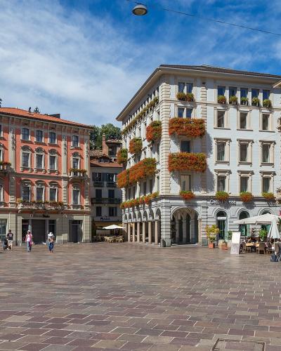 A wide-angle view of a large, paved European city square surrounded by multi-story buildings with pastel facades, flower boxes, and outdoor cafe seating under white umbrellas.