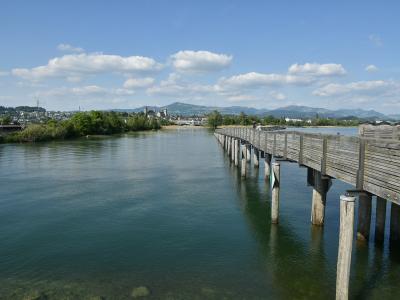 A long wooden footbridge extending across a wide body of water towards a town with a prominent church tower and hills in the background under a partly cloudy sky.