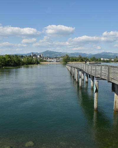 A long wooden footbridge extending across a wide body of water towards a town with a prominent church tower and hills in the background under a partly cloudy sky.