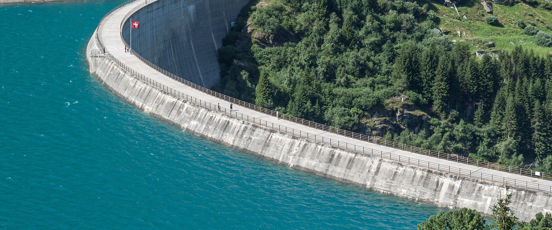 A large concrete arch dam wall curving across a turquoise reservoir, with a Swiss flag flying on the crest and a road running along the top, surrounded by green forested mountains.