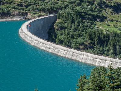 A large concrete arch dam wall curving across a turquoise reservoir, with a Swiss flag flying on the crest and a road running along the top, surrounded by green forested mountains.