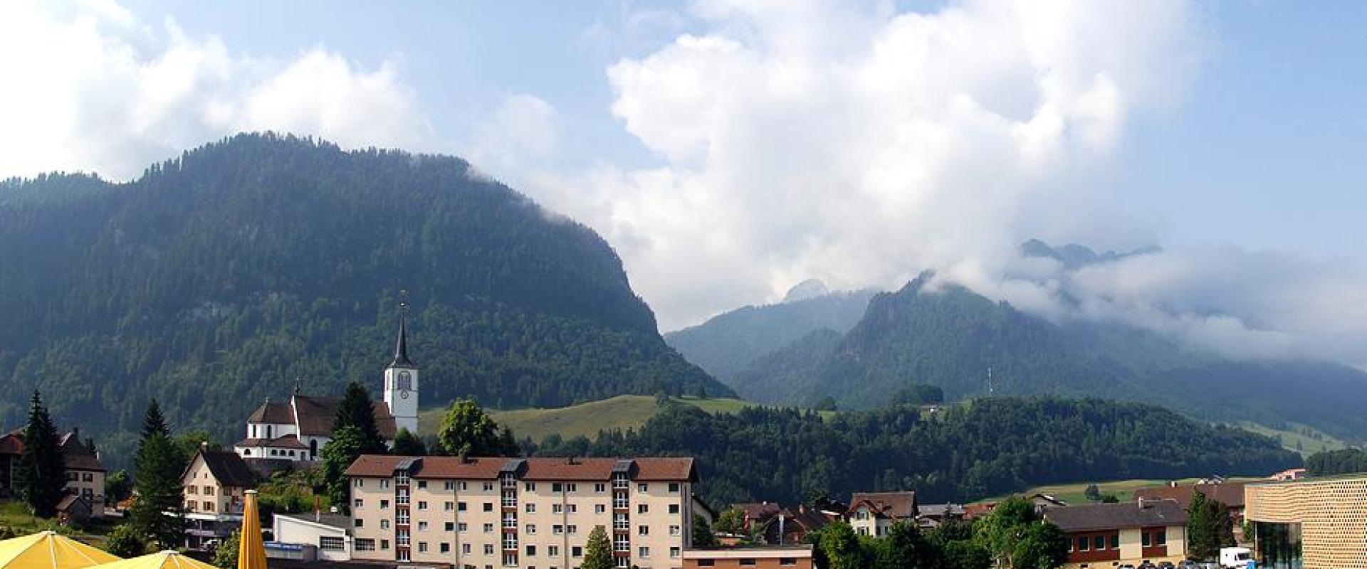 A scenic view of the village of Charmey in Switzerland, featuring a large parking lot, a church with a white steeple, residential buildings, and mountains in the background, with yellow umbrellas in the foreground.