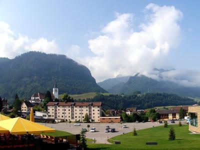 A scenic view of the village of Charmey in Switzerland, featuring a large parking lot, a church with a white steeple, residential buildings, and mountains in the background, with yellow umbrellas in the foreground.