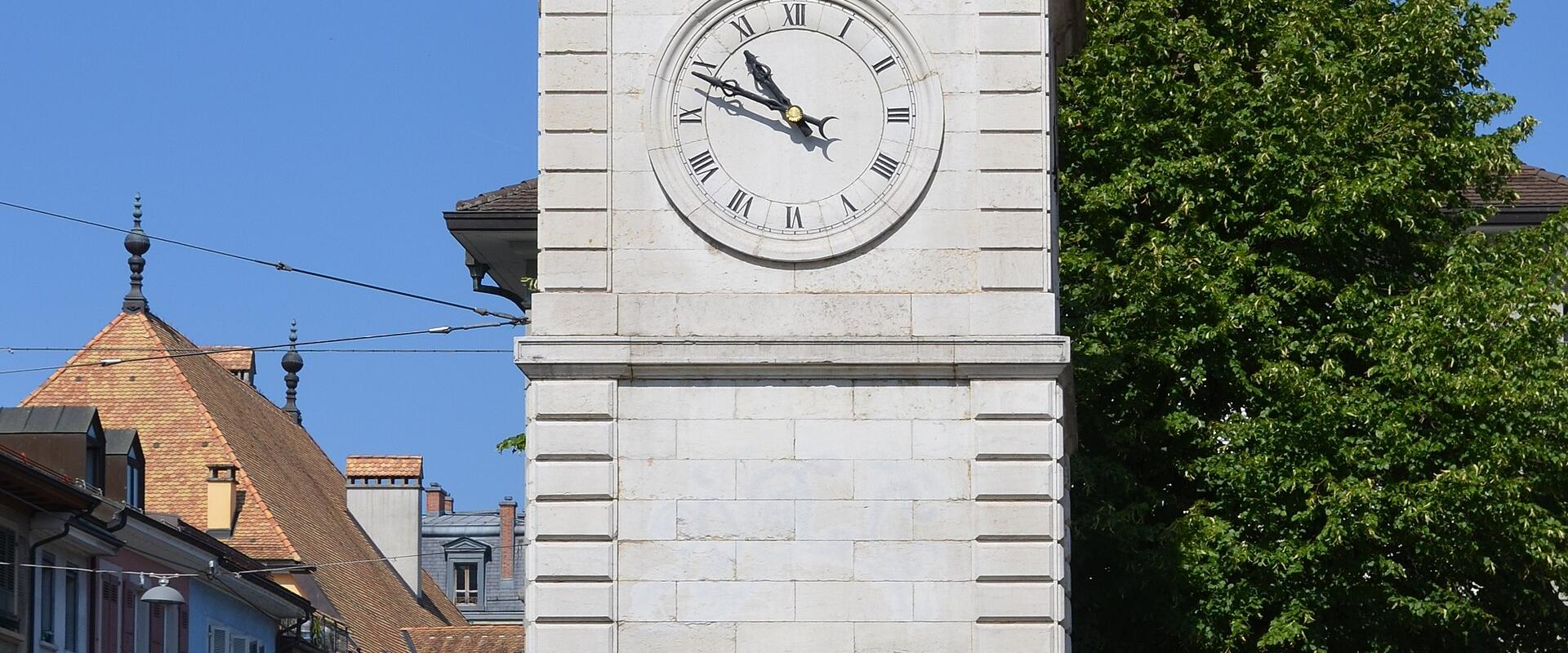 A tall, medieval stone clock tower with a green copper cupola and bell chamber, situated on a street corner next to a cream-colored building with green shutters, under a clear blue sky.