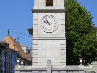 A tall, medieval stone clock tower with a green copper cupola and bell chamber, situated on a street corner next to a cream-colored building with green shutters, under a clear blue sky.