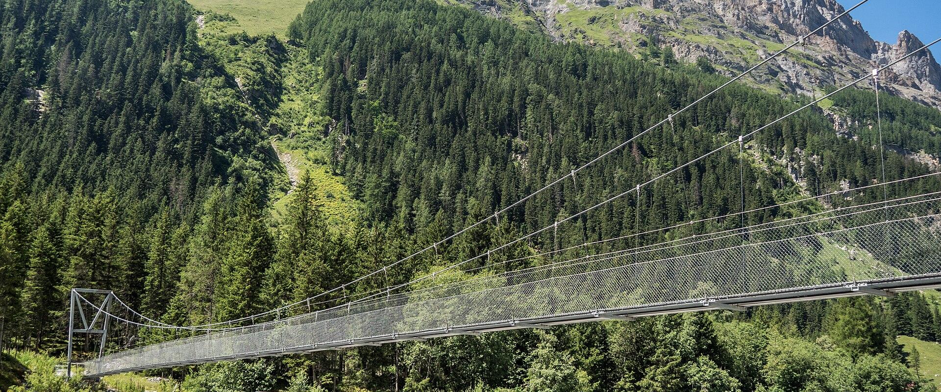 A long metal suspension footbridge spanning a rocky riverbed with a steep, forested mountainside rising in the background.