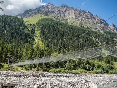 A long metal suspension footbridge spanning a rocky riverbed with a steep, forested mountainside rising in the background.