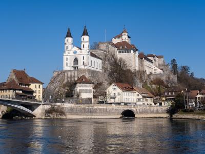 A wide-angle photograph of Aarburg Castle and the adjacent St. Martin's Church perched on a rocky cliff above the Aare River, with a modern bridge crossing the water in the foreground.
