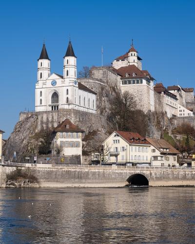 A wide-angle photograph of Aarburg Castle and the adjacent St. Martin's Church perched on a rocky cliff above the Aare River, with a modern bridge crossing the water in the foreground.