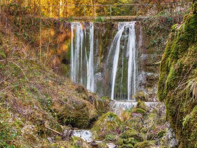 A waterfall cascading over a rocky ledge into a mossy stream bed, with a metal railing visible along the top edge of the cliff.