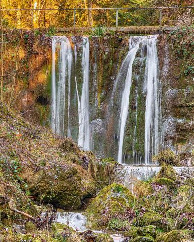 A waterfall cascading over a rocky ledge into a mossy stream bed, with a metal railing visible along the top edge of the cliff.