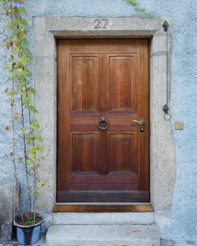 A close-up photograph of a wooden double door set within a stone frame, featuring the number 27 above the lintel, a metal ring knocker, and a climbing vine in a pot to the left.