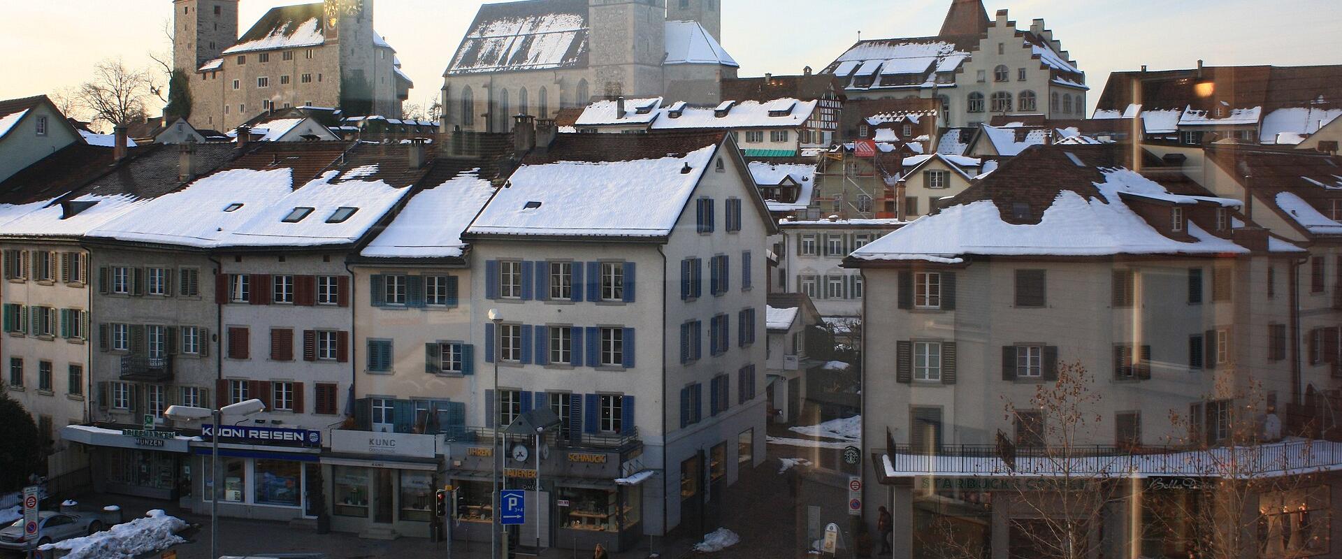 A snowy street scene in a Swiss town featuring a prominent medieval castle with two round towers and a large church with twin towers on a hill behind traditional buildings.