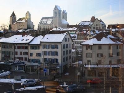 A snowy street scene in a Swiss town featuring a prominent medieval castle with two round towers and a large church with twin towers on a hill behind traditional buildings.