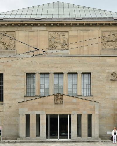 A neoclassical stone building facade featuring three large sculpted reliefs of horses and riders in the upper frieze, a lion sculpture on the right wall, and a large dark bronze gate (the "Hell Gate" by Auguste Rodin) on the far right.