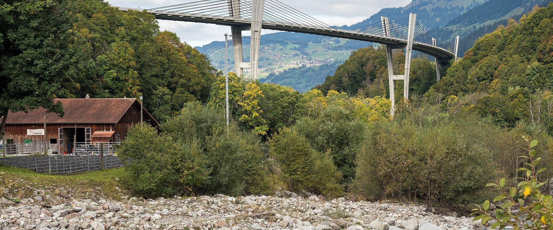 A modern cable-stayed bridge with tall concrete pylons spanning a rocky river valley, with a wooden building and forested hills in the foreground and background.
