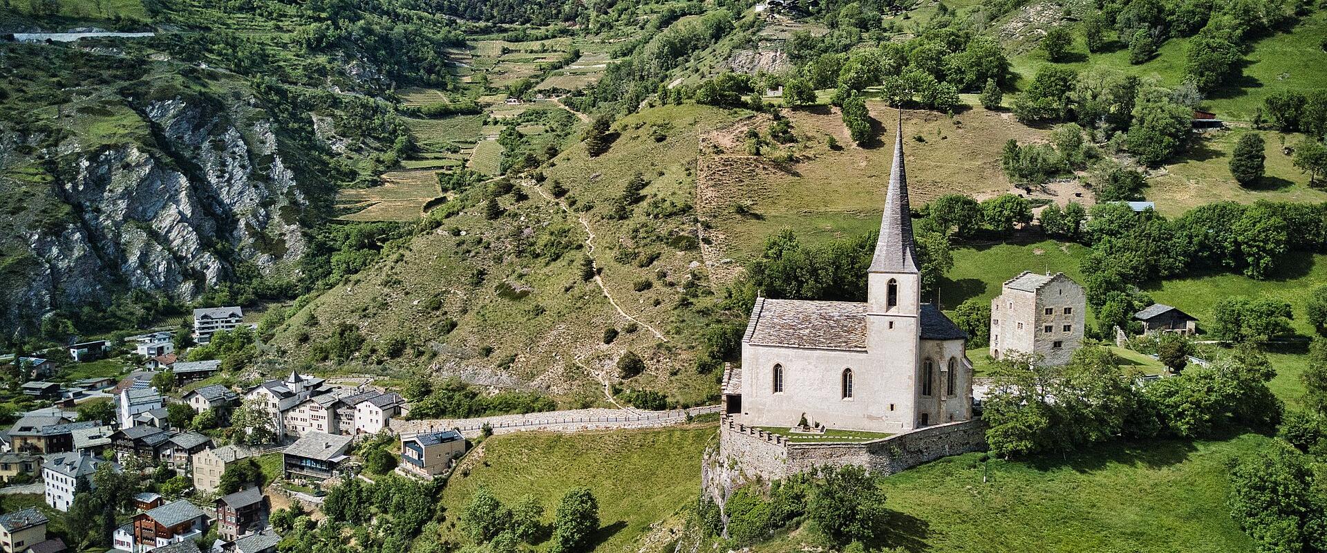 An aerial view of a stone church with a tall, pointed spire situated on a grassy hilltop overlooking a village nestled in a green valley.