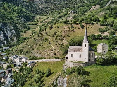 An aerial view of a stone church with a tall, pointed spire situated on a grassy hilltop overlooking a village nestled in a green valley.