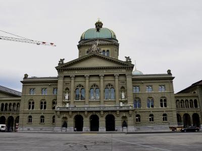 A large, ornate neoclassical government building with a prominent central dome and pediment inscription reading 'CURIA CONFŒDERATIONIS HELVETICAE', situated in a wide paved plaza under an overcast sky.