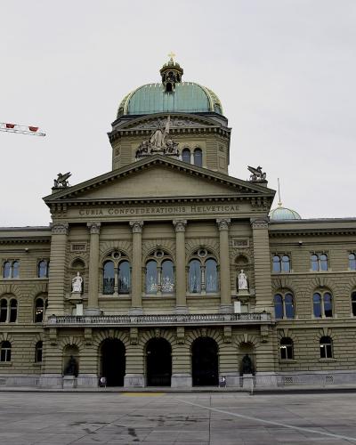 A large, ornate neoclassical government building with a prominent central dome and pediment inscription reading 'CURIA CONFŒDERATIONIS HELVETICAE', situated in a wide paved plaza under an overcast sky.