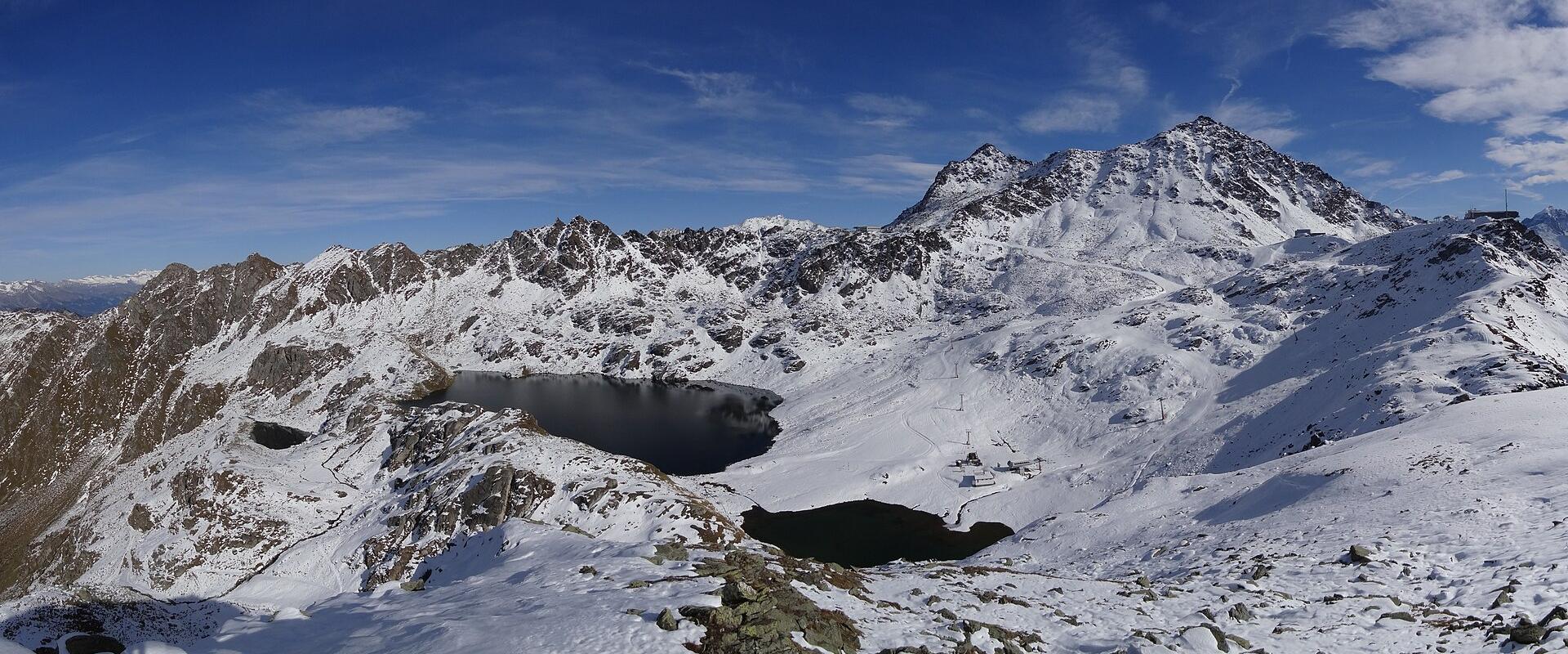 A high-altitude alpine landscape featuring two dark lakes nestled among snow-covered rocky peaks under a blue sky, with a cable car station visible on the ridge to the right.