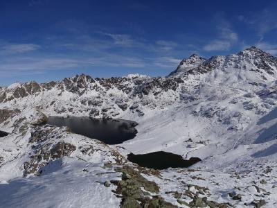 A high-altitude alpine landscape featuring two dark lakes nestled among snow-covered rocky peaks under a blue sky, with a cable car station visible on the ridge to the right.