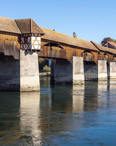 A long, covered wooden bridge with a tiled roof and stone piers spanning a wide river, with buildings visible on the far bank.