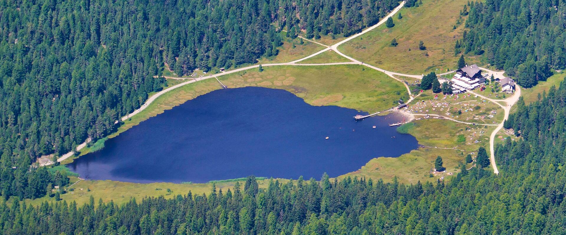 An aerial view of a dark blue alpine lake surrounded by dense coniferous forest and green meadows, with a large building complex and dirt roads visible on the eastern shore.