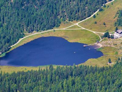 An aerial view of a dark blue alpine lake surrounded by dense coniferous forest and green meadows, with a large building complex and dirt roads visible on the eastern shore.