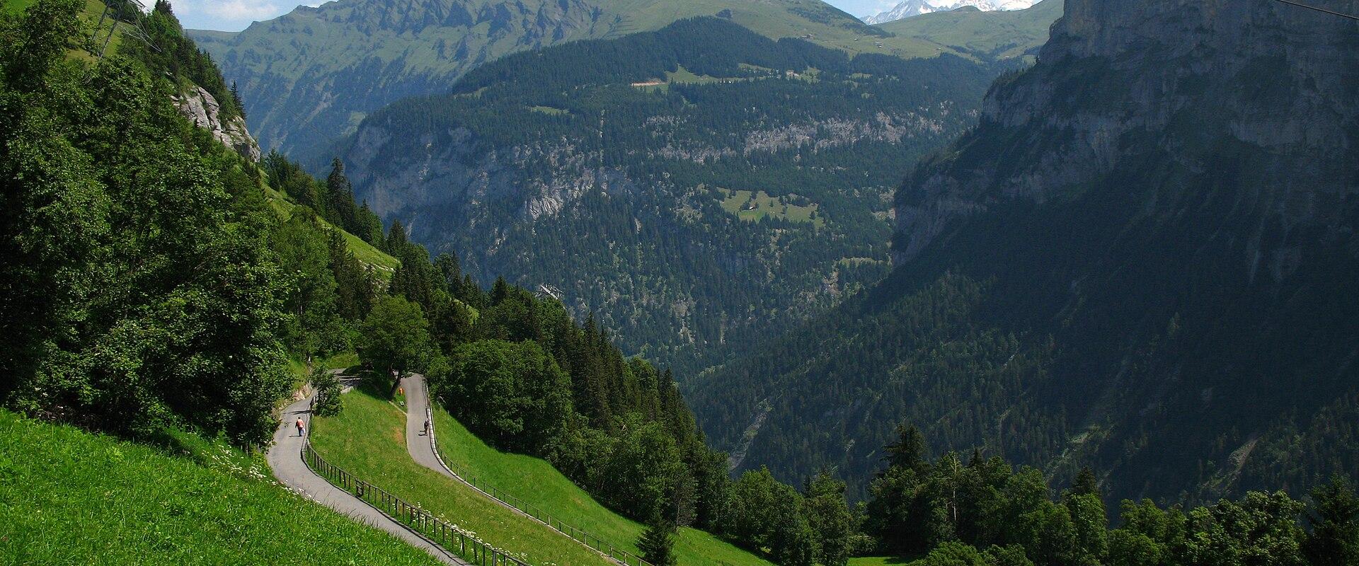 A winding paved road curves along a steep, grassy mountainside with a small wooden hut in the foreground and distant alpine peaks under a blue sky.