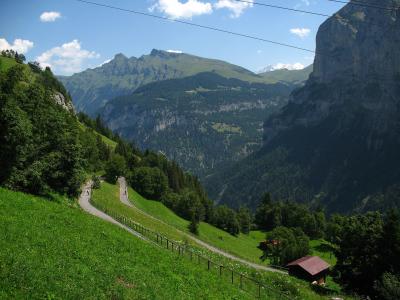 A winding paved road curves along a steep, grassy mountainside with a small wooden hut in the foreground and distant alpine peaks under a blue sky.