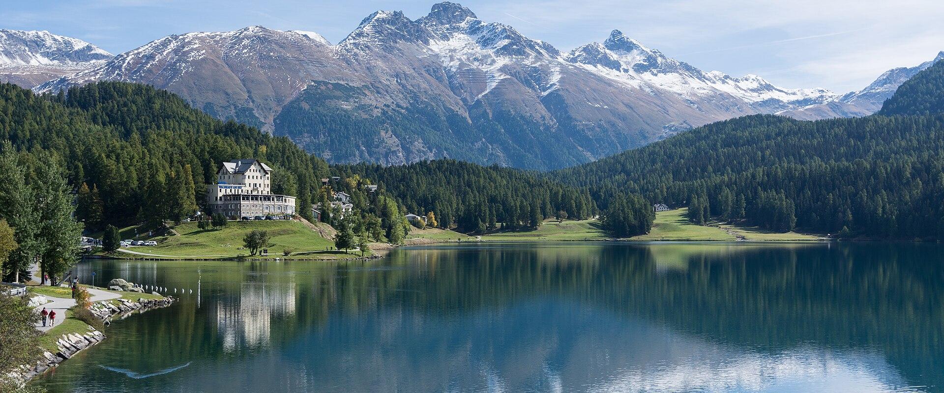 A scenic view of a large alpine lake with a prominent white hotel building on the left shore, surrounded by green forests and snow-capped mountains in the background.