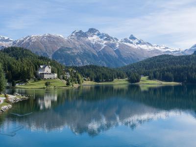 A scenic view of a large alpine lake with a prominent white hotel building on the left shore, surrounded by green forests and snow-capped mountains in the background.