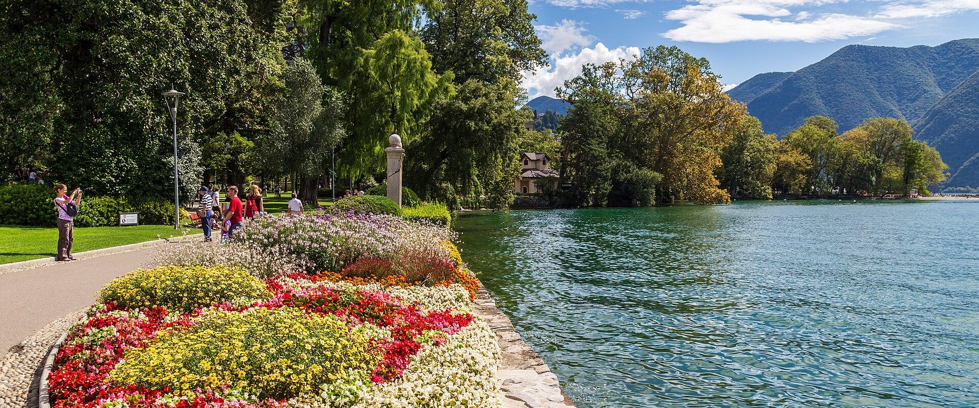 A paved promenade along a lake shore lined with vibrant, multi-colored flowerbeds, trees, and people walking, with mountains in the background.