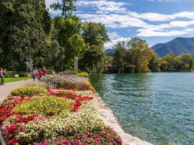 A paved promenade along a lake shore lined with vibrant, multi-colored flowerbeds, trees, and people walking, with mountains in the background.