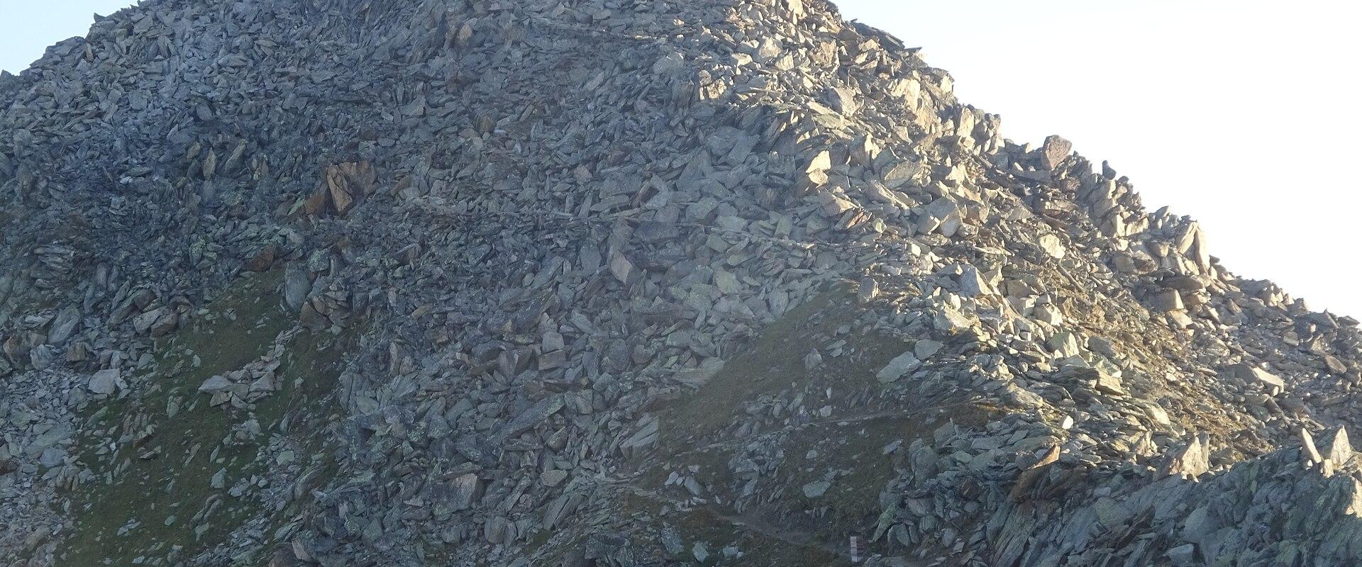 A close-up view of a steep, rocky mountain summit with a small cross at the peak and a narrow trail winding up the scree slope.