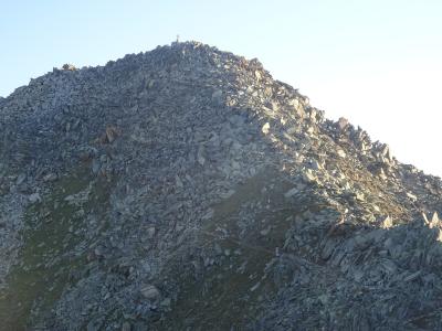 A close-up view of a steep, rocky mountain summit with a small cross at the peak and a narrow trail winding up the scree slope.