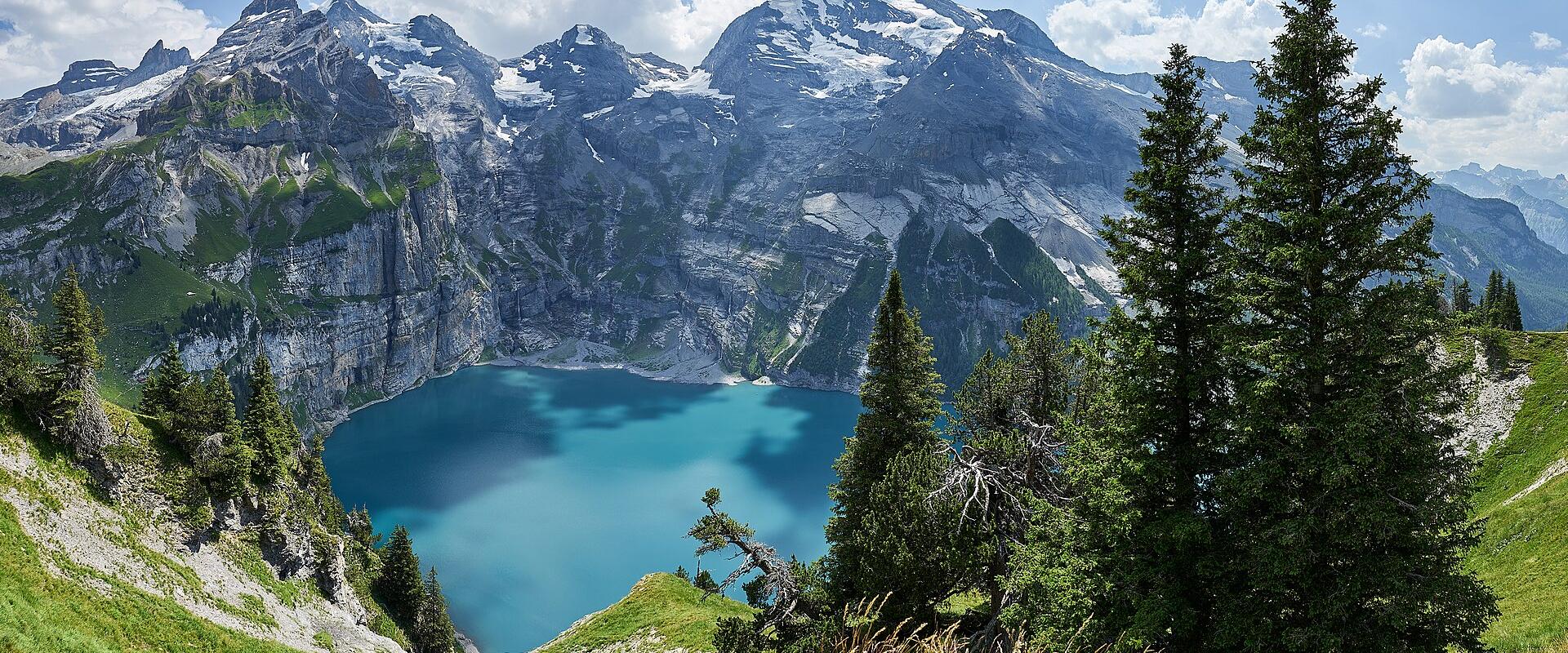 A panoramic view of a turquoise alpine lake surrounded by steep, rocky mountain slopes with snow patches and evergreen trees in the foreground.