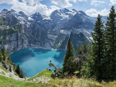 A panoramic view of a turquoise alpine lake surrounded by steep, rocky mountain slopes with snow patches and evergreen trees in the foreground.