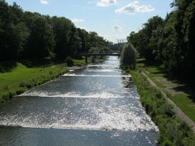 A wide, shallow river flows through a green park with trees lining both banks, a bridge in the distance, and people relaxing on the grass; the filename is 'Fluß Wiese flussabwärts Lange Erlen'.