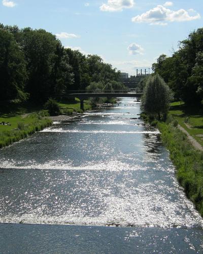A wide, shallow river flows through a green park with trees lining both banks, a bridge in the distance, and people relaxing on the grass; the filename is 'Fluß Wiese flussabwärts Lange Erlen'.