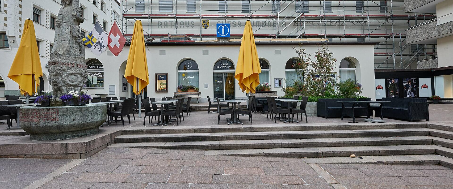A paved town square featuring a stone fountain with a statue on the left, yellow umbrellas, and a white building in the background displaying the text "RATHAUS" and a Swiss flag.