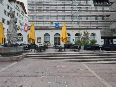 A paved town square featuring a stone fountain with a statue on the left, yellow umbrellas, and a white building in the background displaying the text "RATHAUS" and a Swiss flag.
