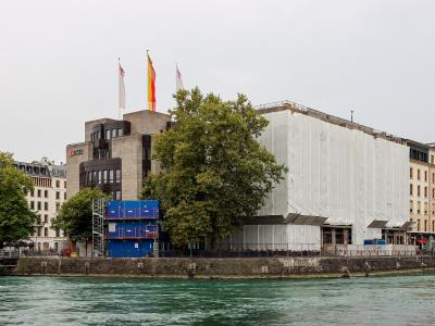 A view of the Quai des Bergues in Geneva, Switzerland, showing the Pont de la Machine bridge over the Rhône river with buildings featuring "VACHERON CONSTANTIN" and "ZENITH" signage on the right.