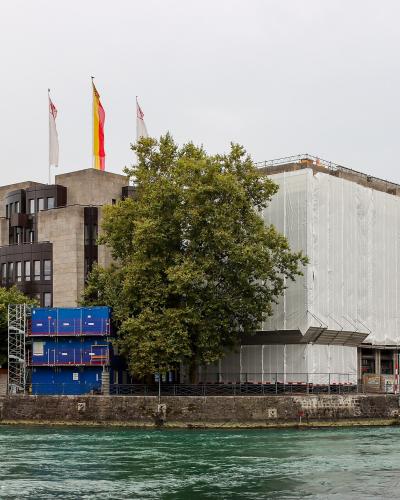 A view of the Quai des Bergues in Geneva, Switzerland, showing the Pont de la Machine bridge over the Rhône river with buildings featuring "VACHERON CONSTANTIN" and "ZENITH" signage on the right.