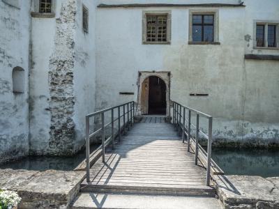 A wooden footbridge with metal railings leads to an arched doorway in the white-washed wall of a historic building situated over a narrow canal or moat.