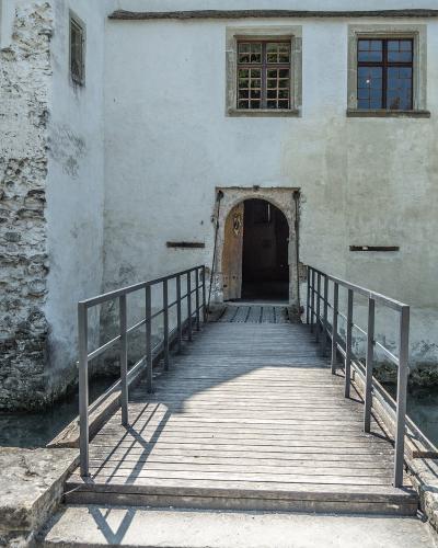 A wooden footbridge with metal railings leads to an arched doorway in the white-washed wall of a historic building situated over a narrow canal or moat.