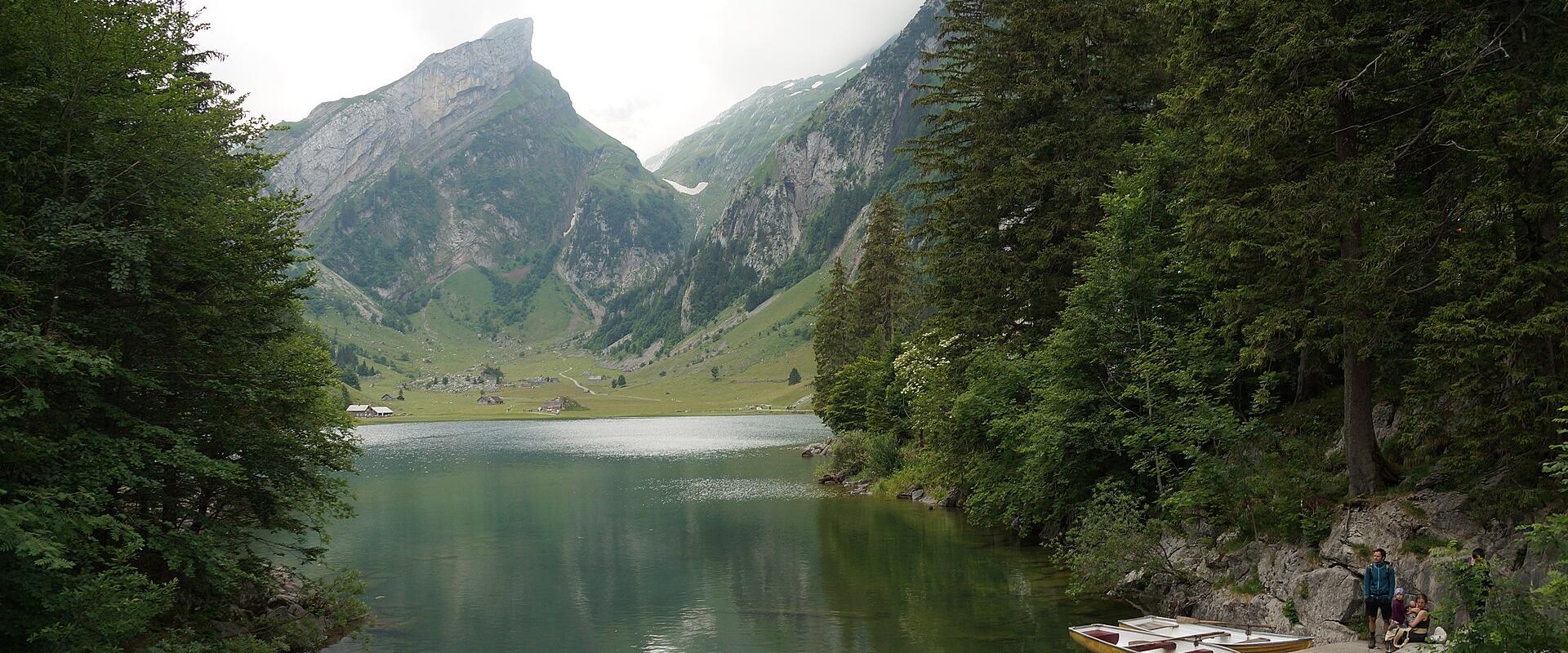 A scenic view of a lake surrounded by green forests and mountains, with a distinctive pointed mountain peak in the background and yellow rowboats on the shore.