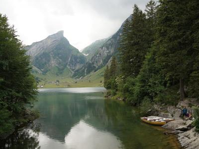 A scenic view of a lake surrounded by green forests and mountains, with a distinctive pointed mountain peak in the background and yellow rowboats on the shore.